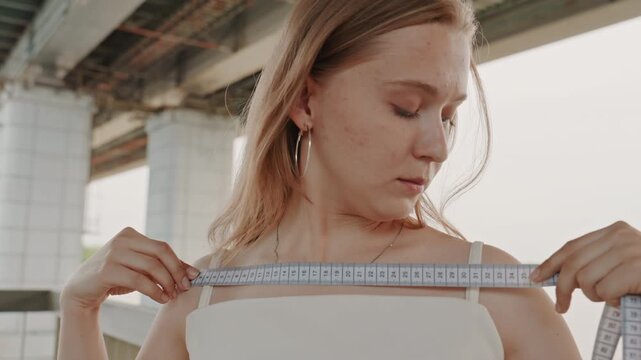 Woman measures her bust with tape measure under overpass wearing white top and hoop earrings. background features structural columns of bridge and soft daylight lighting creating calm
