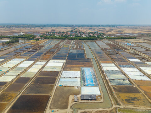 Aerial view of the Salt Ponds featuring geometric salt evaporation pans with varying water colors, surrounded by rural landscape and a small village in Kedung, Central Java, Indonesia.