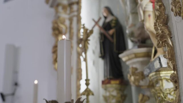 A tall candle on a chapel altar beside gilded carving, with the statue of a nun holding a crucifix out of focus behind.