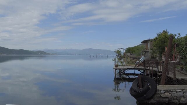 Lake Ohrid at Lin in Albania, flat water reflecting clouds and hills, beside an old wooden pier with a tire fender on the stone shore.