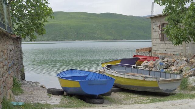 Painted rowboats pulled up on the shore of Lake Ohrid in the village of Lin, Albania, beside a block house, rocks and old tires.