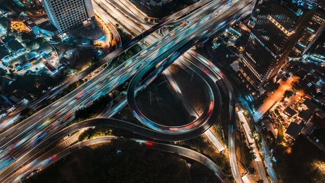 Aerial view of a multi-level highway interchange at night with light trails from moving vehicles, surrounded by illuminated skyscrapers in Jakarta, Indonesia.