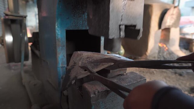 Close-up of a blacksmith using tongs to hold a metal workpiece on an anvil while a heavy forging hammer is positioned above it. A traditional forge and a burning fire are visible in the background.
