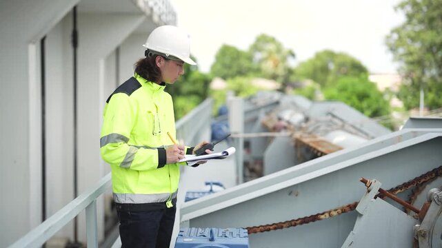Engineer examines floodgate equipment while taking notes during field inspection, documenting machinery condition, maintenance needs and operational status at water management facility.