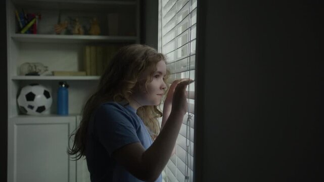 Child thinking quietly by the window. Kid looking outside deep in thought think. Thoughtful child near window blinds. Thinking kid at home. Child lost in thinking in soft window light. Kids think.