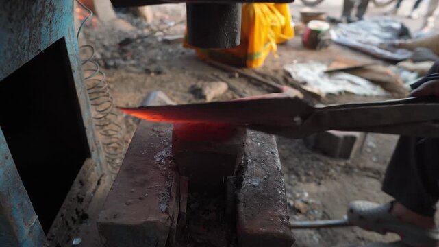 Close-up of a blacksmith using tongs to hold heated metal on an anvil while a mechanical power hammer shapes the workpiece in an active metal forging workshop.