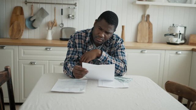 Financial troubles and utility bills, man looking at money and papers at home. Poverty and jobless. Portrait of african male person sitting alone on kitchen and thinking about budget and payments