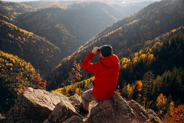 Fototapeta premium Man sitting on a rocky ledge and looking through binoculars at an autumn mountain landscape.