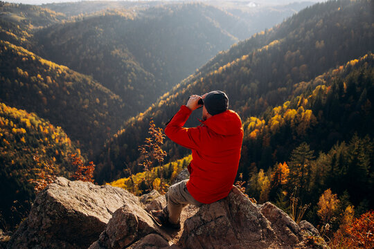 Man sitting on a rocky ledge and looking through binoculars at an autumn mountain landscape.