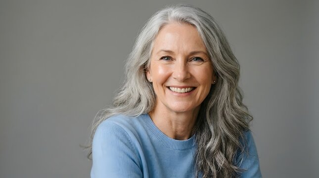 Beautiful smiling senior woman with natural long silver hair looking at camera, confident mature lady in blue sweater posing over gray background, healthy aging and lifestyle concept.