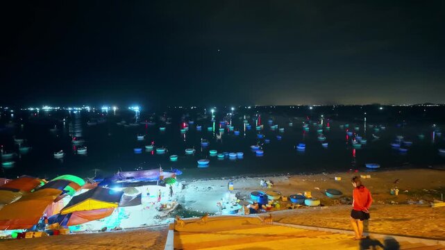 Timelapse of nighttime view of Mui Ne fishing village in Vietnam, featuring numerous fishing boats and colorful basket boats floating on the dark sea. 