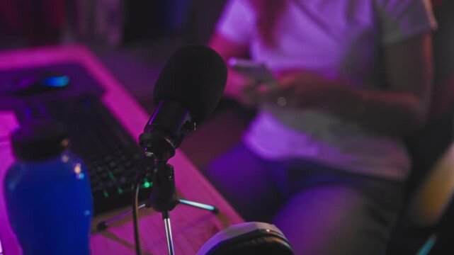 Woman scrolling smartphone with hands visible, seated in gaming chair by neon controller sign, microphone and keyboard visible in streaming studio; calm focus.