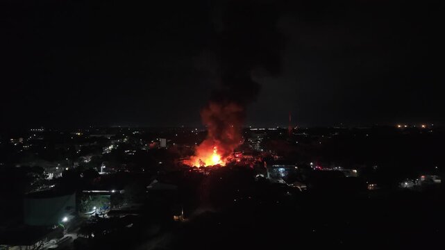 Philippines, Palawan, Puerto Princesa 10.05.2026: Aerial night view of a residential fire in Barangay Masipag sending a black smoke column above city lights, with a lattice tower on the skyline.