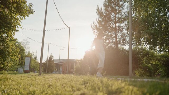 Woman in white dress walks and dances outdoors on grass holding object like headphones or equipment. scene features sunlight backlighting her figure with trees string lights poles greenery