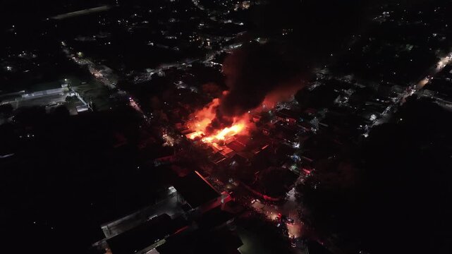 Philippines, Palawan, Puerto Princesa 10.05.2026: Aerial night view of a water jet aimed at burning houses in Barangay Masipag, with a fire truck, people on the street and thick smoke nearby.