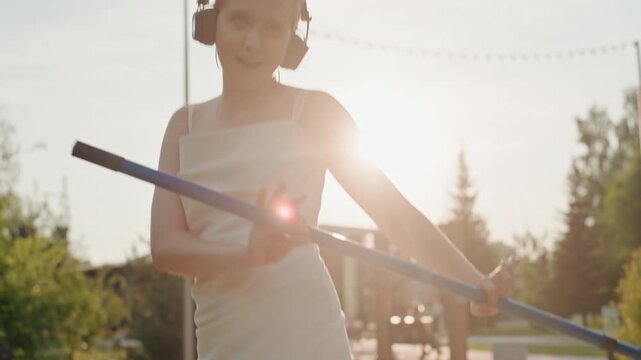 Young woman wearing headphones practices pole vaulting in park during sunset. Woman holds and maneuvers blue padded bar preparing for athletic event with focused movements against outdoor