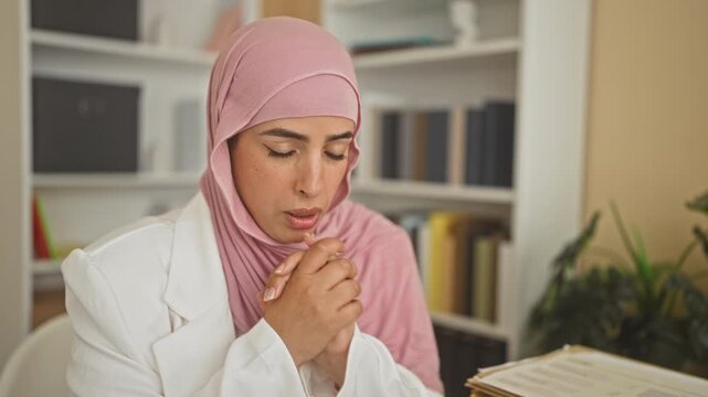Woman in pink hijab and white blazer clasping hands near mouth over folders and documents on a desk in an office building; concern reflection.