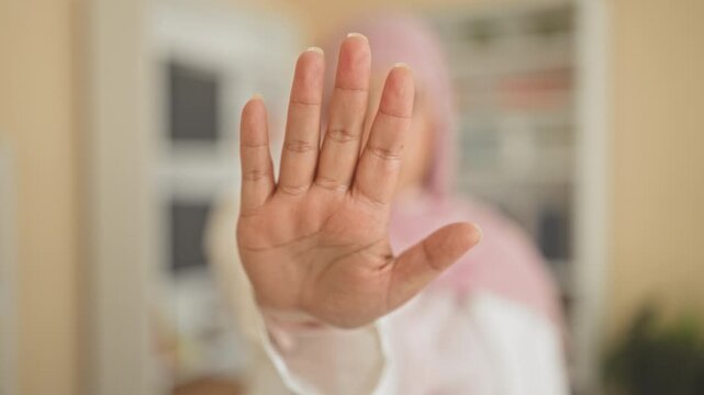Woman palm forward stop gesture toward camera in studio, hijab wearing young arab with bookshelf and plant visible; privacy refusal.