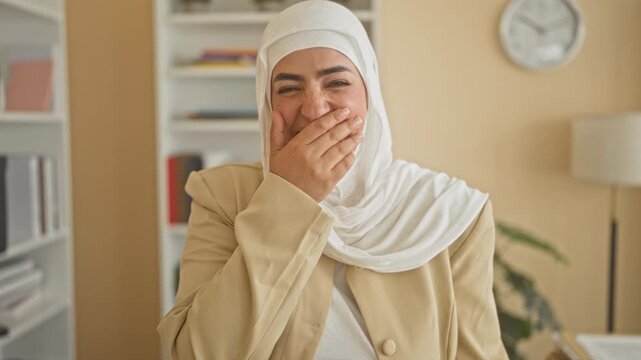 Woman covering mouth with hand laughing in a building office with bookshelf and clock visible; joy warmth connection.