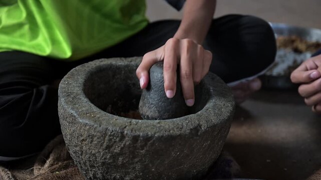Close-up video footage of a person's hands using a traditional stone mortar and pestle (okhli) to grind spices by hand, a time-honored method still practiced in rural Indian households.