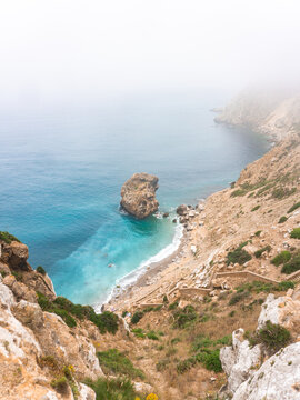 Rocky coastline overlooking a turquoise sea with distant sea stack in heavy mist