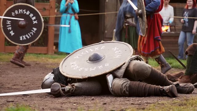 Two Viking warriors fighting with swords and shields while fallen warrior lies in foreground.