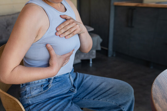 Unrecognizable woman performing a breast self-examination while sitting in an armchair at home. Breast pain, breast cancer, mastopathy, early detection of a breast tumor. Close-up of hands.