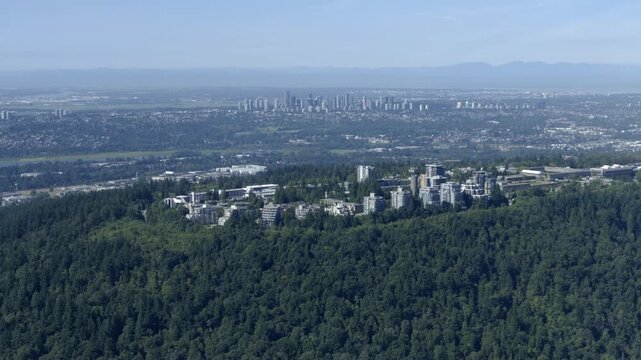 Simon Fraser University Atop Forested Burnaby Mountain With Metro Vancouver Downtown Skyline In The Background In Canada. - aerial shot