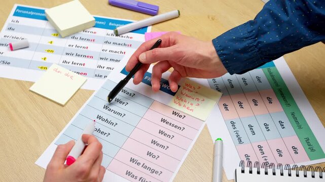 Hands point with a pen at printed German grammar tables and words on a wooden desk. A foreign language education and learning concept.