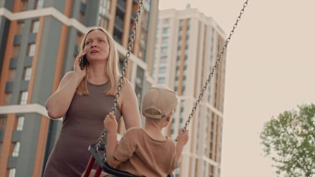 Woman talks on her phone while standing next to young boy swinging in outdoor playground with tall buildings in background. scene shows soft natural light and calm interaction between mother