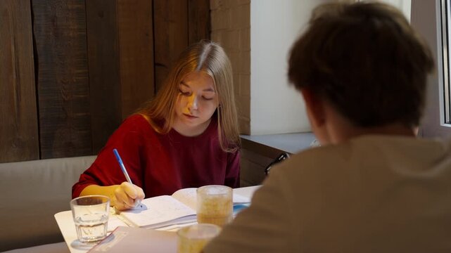Teen girl writing homework with classmate at cafe table
