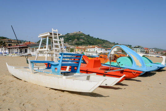 Colorful pedal boats and a lifeguard chair on the sandy beach at Silvi, with the hilltop old town in the background under a clear blue sky.