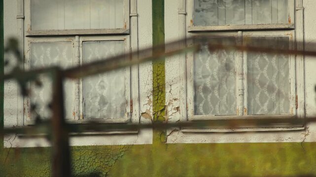 A rustic view of two old wooden windows painted white on a weathered green house. The paint is aged, cracked, and peeling, revealing the passage of time. The windows are seen through a wooden fence.
