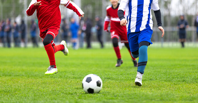 Youth Soccer Tournament Action. Junior Football Player in Red Kit Running Towards Soccer Ball Player in Blue and White Striped Kit. Kids Playing Soccer Game on Grass Pitch