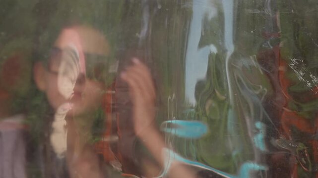 Young woman adjusts her sunglasses and face mask through glass window with reflections of abstract patterns visible. Woman moves slightly while interacting with transparent surface
