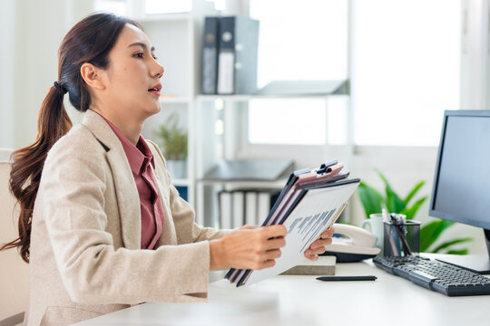A serious Asian businesswoman is intently searching through a stack of documents while sitting at her desk in her office.