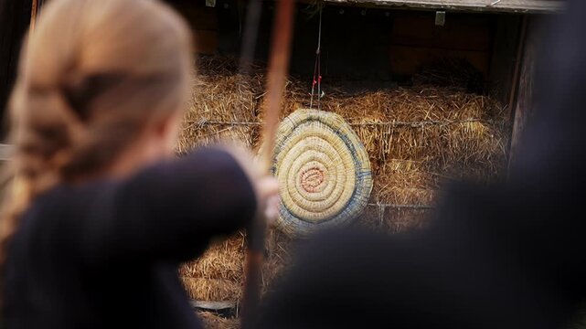 Viking woman aiming arrow with bow at straw target during training outdoors.