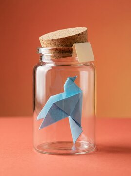 Blue origami bird inside glass jar with cork lid on orange backdrop
