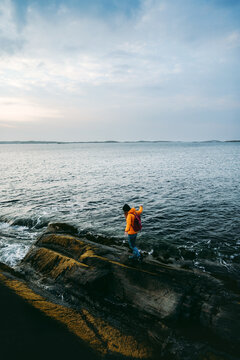 Traveler woman in orange jacket travel with backpack walking by sea on rocky coast in Norway outdoor, active vacations solo eco tourism adventure freedom getaway active lifestyle