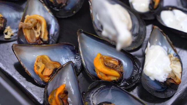 A spoon drizzles thick garlic cream sauce onto orange mussel meat resting in glossy blue black half shells set in a dark tray, an appetizer being finished before baking.