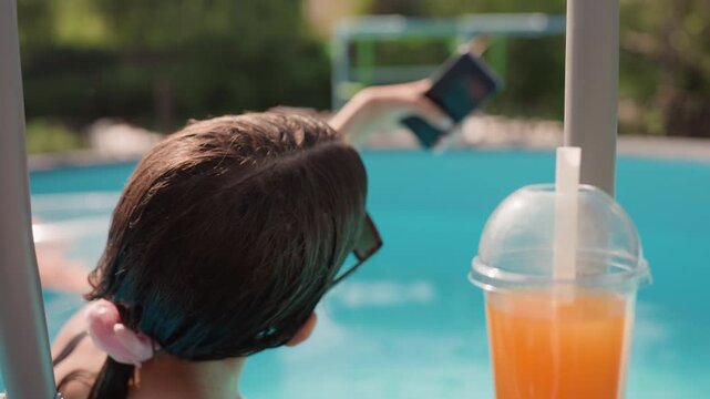 Young woman relaxes in pool holding her smartphone to take photos or. Woman wears sunglasses and has wet hair tied with pink scrunchie enjoying sunny outdoor setting by water. plastic cup