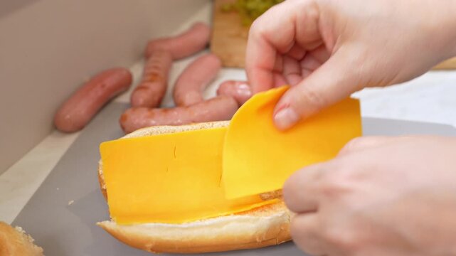 Human hands layer yellow cheddar slices into a soft bun, then tuck a cooked sausage inside on a gray cutting board, with relish and more sausages waiting nearby in a home kitchen.