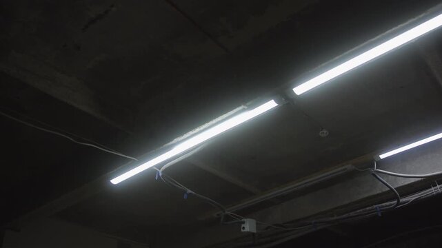 Diagonal fluorescent tube lights glowing under rough concrete ceiling with exposed conduit, junction box, and hanging cables, from low-angle wide viewpoint in industrial interior