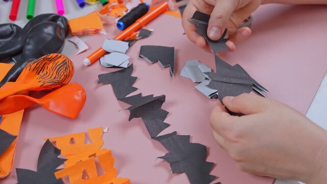 Female hands lay out black paper bat cutouts in neat rows beside scraps, thread and a balloon on a pink surface, a step by step home preparation of seasonal wall decor.
