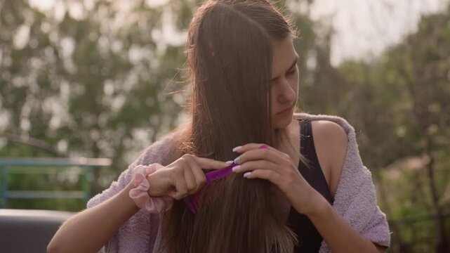 Young woman combs her long brown hair with purple comb in outdoor setting. Woman wears black top and has pink scrunchies on both wrists while soft sunlight illuminates scene against green