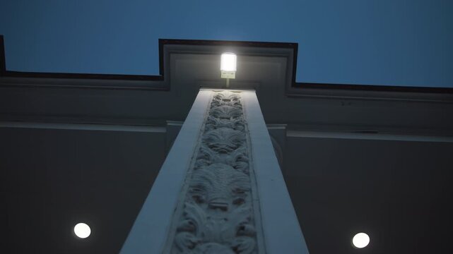 Low-angle view of ornate carved pilaster rising to glowing soffit light beneath stepped cornice on symmetrical facade at twilight