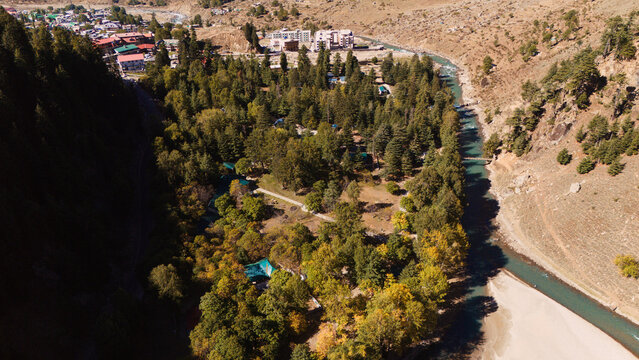 High angle view showcasing lush green conifer trees, riverbanks, and wooden resort cabins nestled in the mountain wilderness of Northern Pakistan.