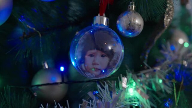 A clear Christmas bauble holding a toddler's photo hangs on a decorated tree among silver ornaments, tinsel and colorful string lights