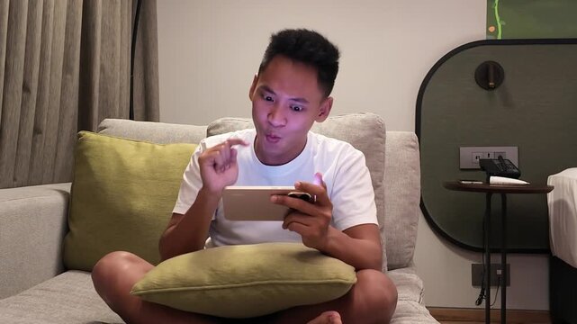 Young Asian man sits on a couch at home, reacting with excitement while playing a mobile game on smartphone late at night. casual, relaxed mood capturing joy, surprise and digital entertainment.