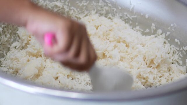 4K Chef hand holding white paddle to fluff cooked Jasmine rice inside large electric rice cooker pot, food preparation process for Asian cuisine.
